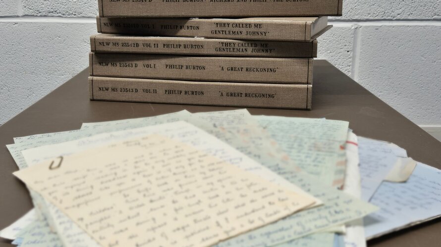 A stack of bound manuscripts with brown covers labelled 'Philip Burton' placed on a table with a pile of papers in view in the foreground